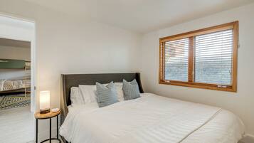 A neatly made bed with white bedding and striped pillows, a bedside table with a lamp, and a window with blinds.