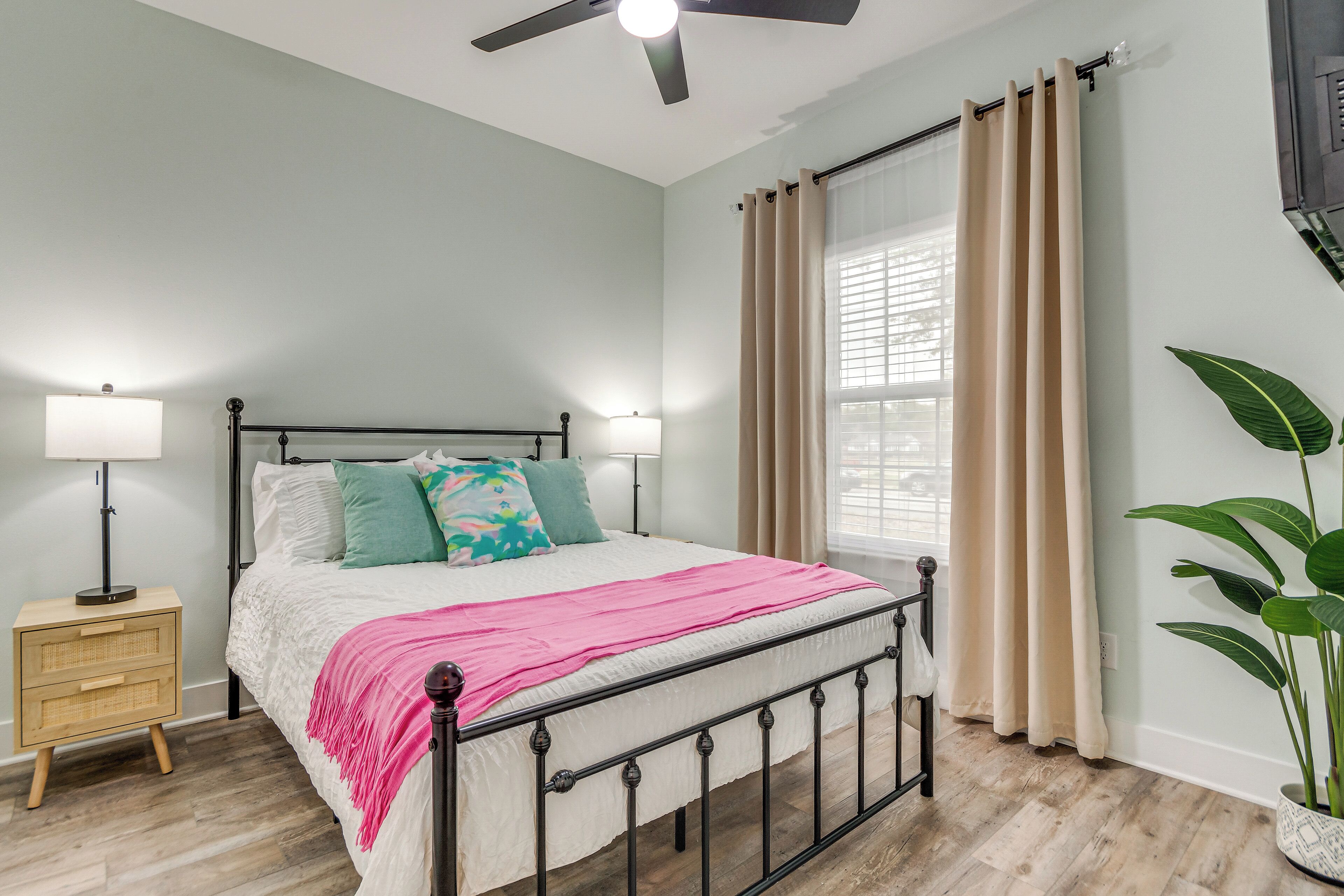 A bedroom with a bed, bedside lamps, a nightstand, a ceiling fan, a window with curtains, and a potted plant.