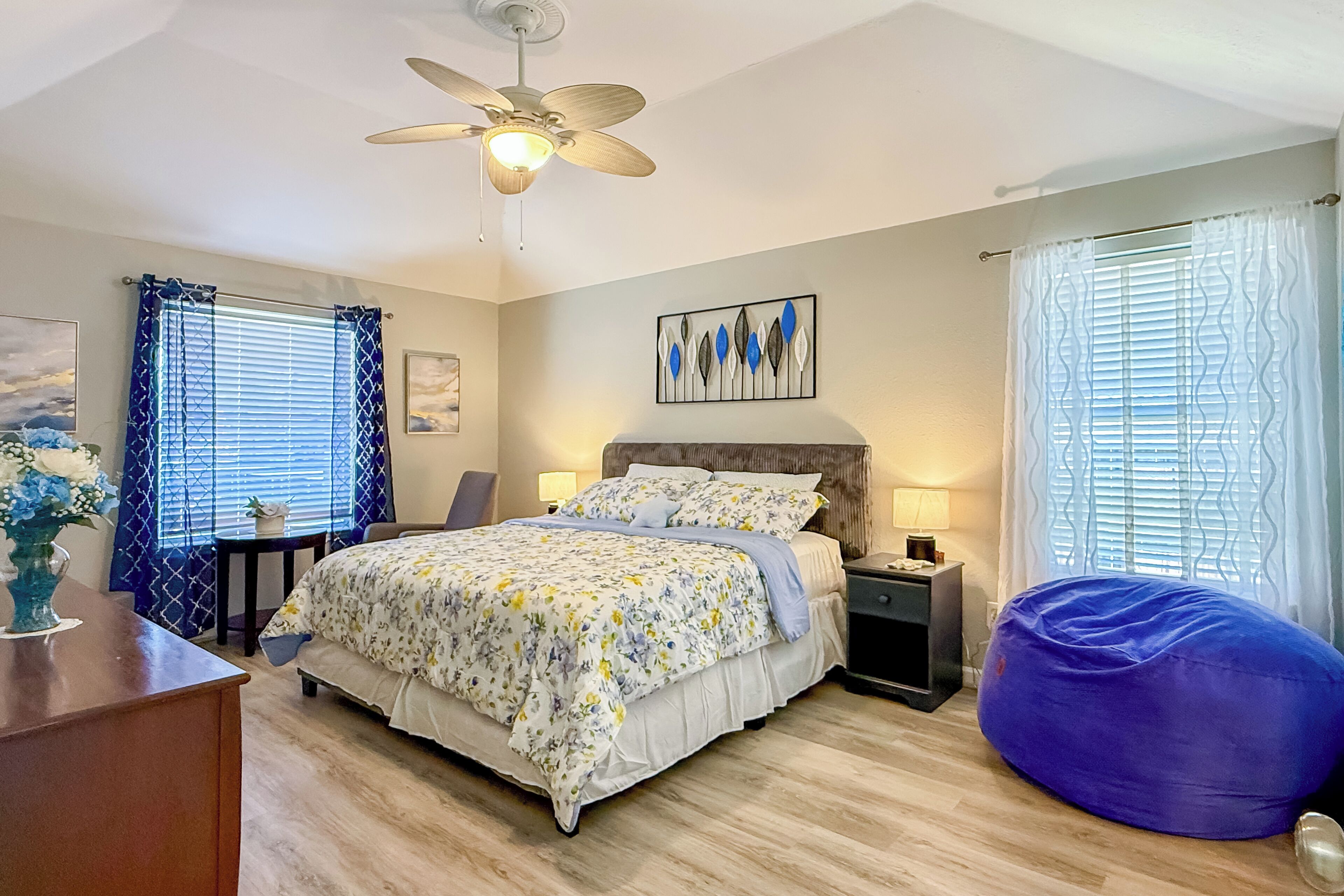 A bedroom with a bed, a ceiling fan, a nightstand, a chair, a bean bag, and a window with curtains.