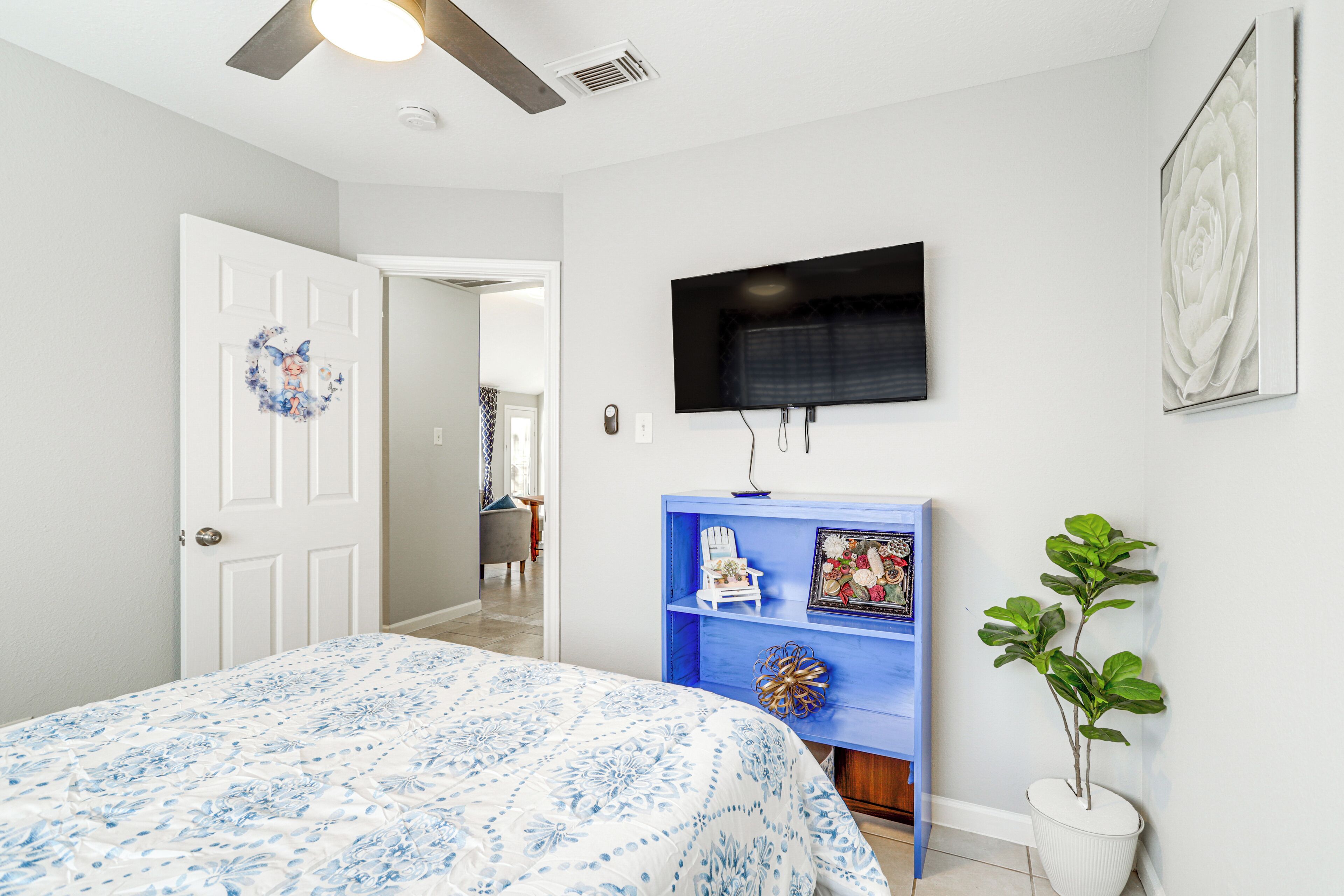 A bedroom with a bed, a television mounted on the wall, a blue shelf with decorative items, and a potted plant.