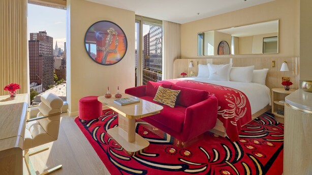 A hotel room with a large bed, a red sofa, a coffee table, a desk, and a view of the cityscape through the window. - Faena New York (New York)