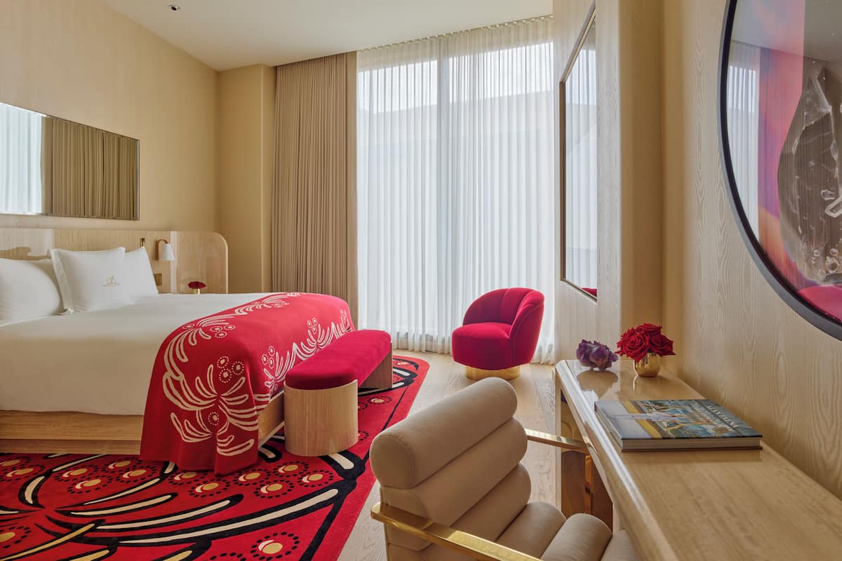 Una habitación de hotel con una cama, un sillón rojo, una mesita con un libro y un espejo grande.