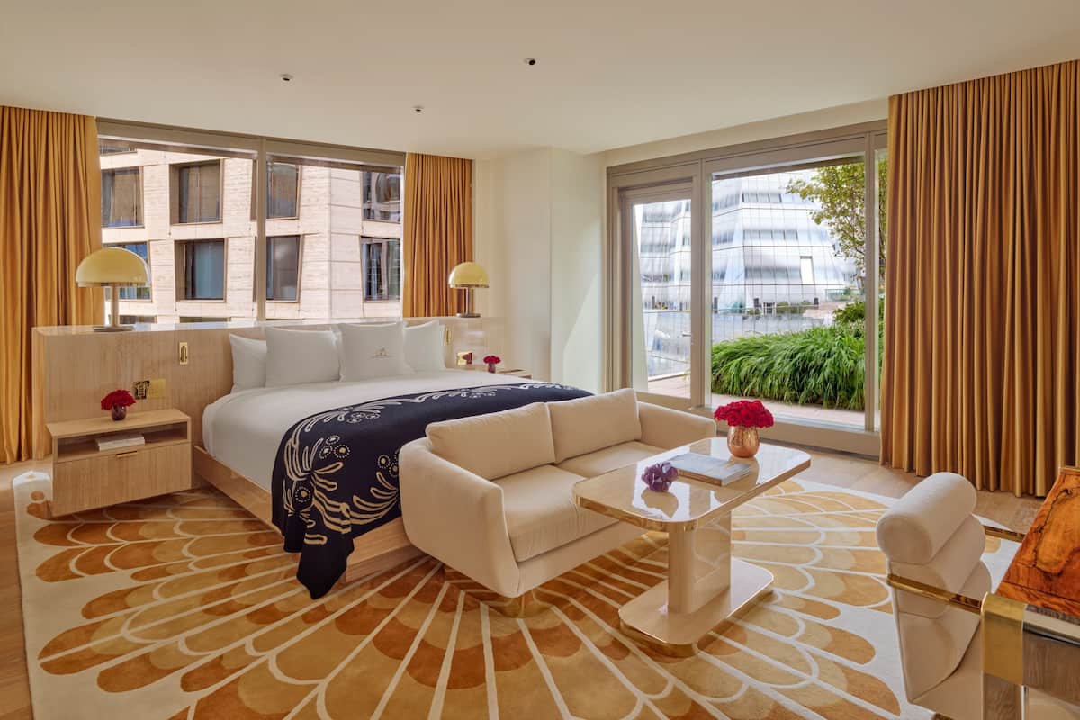 Habitación de hotel moderna con una cama grande, un sofá, una mesa de centro y vistas a la ciudad a través de amplios ventanales.