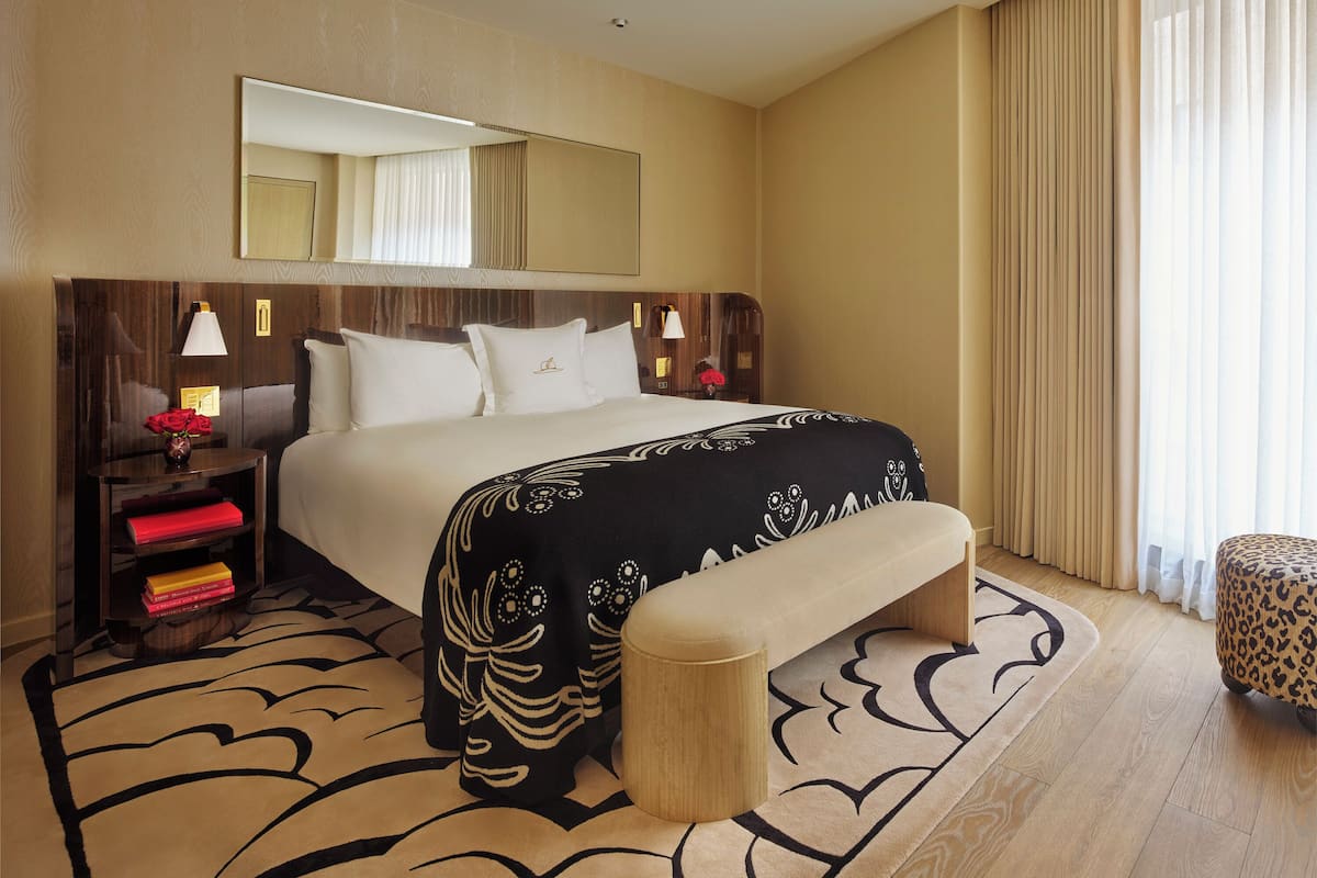 Habitación de hotel con una cama grande, mesitas de noche con lámparas, un espejo sobre la cama, un banco y una ventana con cortinas.