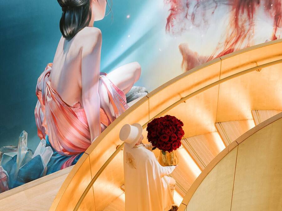 Una persona amb una bata blanca sosté un ram d'roses vermelles en una escala rol·la mentre al fons hi ha un mural d'una dona i un àngel.