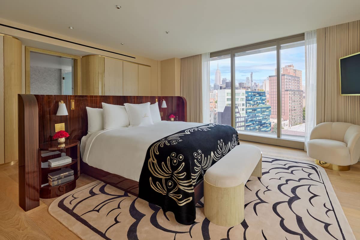 Habitación de hotel con una cama grande, una mesita de noche con lámpara, un taburete pequeño y una vista panorámica de la ciudad a través de amplios ventanales.