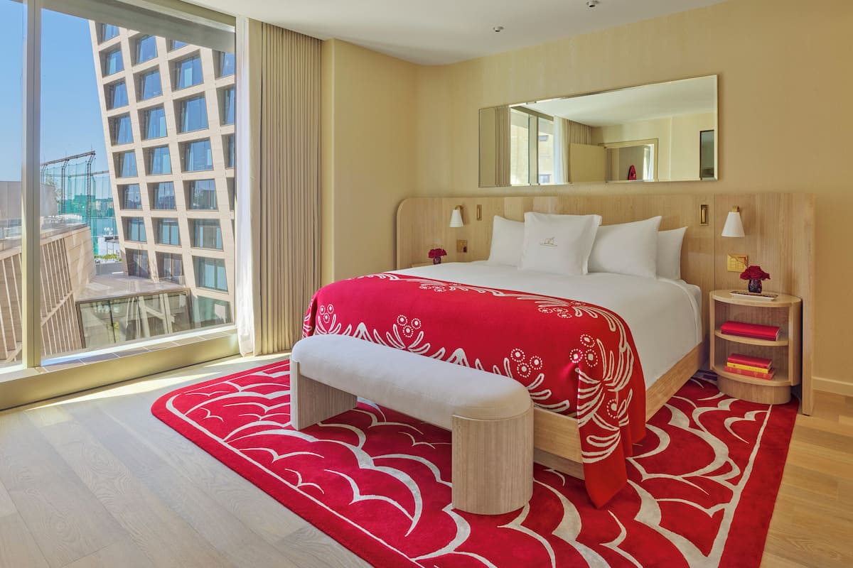 Habitación de hotel con una cama grande, una banca, una mesita de noche con lámpara, un espejo y vista a un edificio.