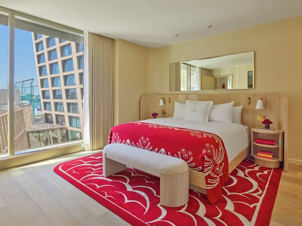 Habitación de hotel con una cama grande, una banca, una mesita de noche con lámpara, un espejo y vista a un edificio.