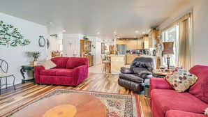 Un vaste espace de vie comprenant un canapé rouge, un fauteuil en cuir et une table basse en bois. On aperçoit une cuisine à l’arrière-plan, avec des meubles de cuisine en bois et un réfrigérateur.