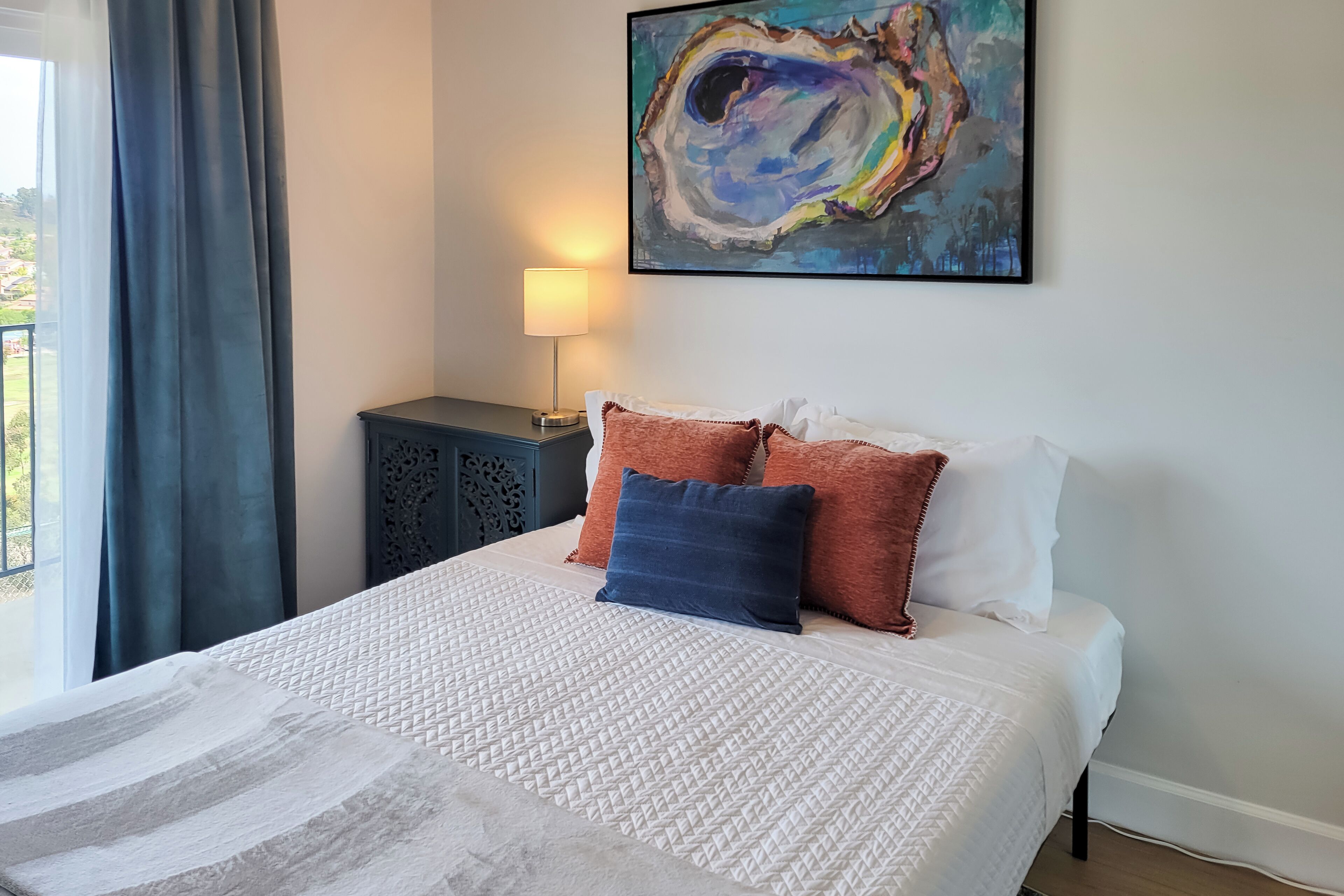 Een opgemaakt bed met een geborduurde spreideken en kussens, een schilderij aan de muur en een lamp op een nachtkastje.