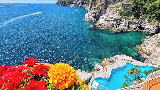 Une vue sur la côte, avec une mer bleue et limpide, une falaise rocheuse et une piscine dotée d’un escalier menant à un niveau supérieur.