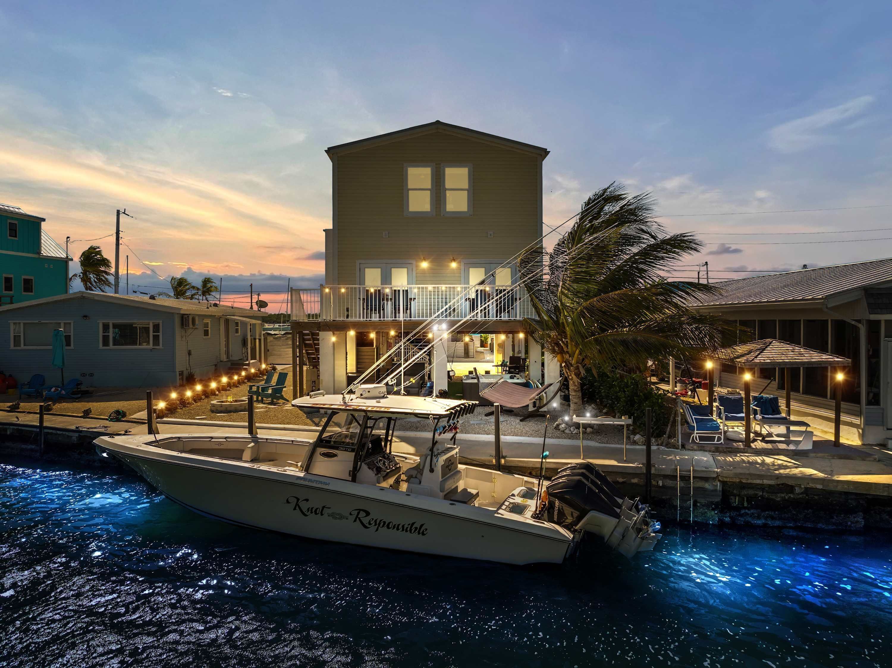 A waterfront property with a boat docked in front, a multi-level house with outdoor seating, and a view of the ocean.