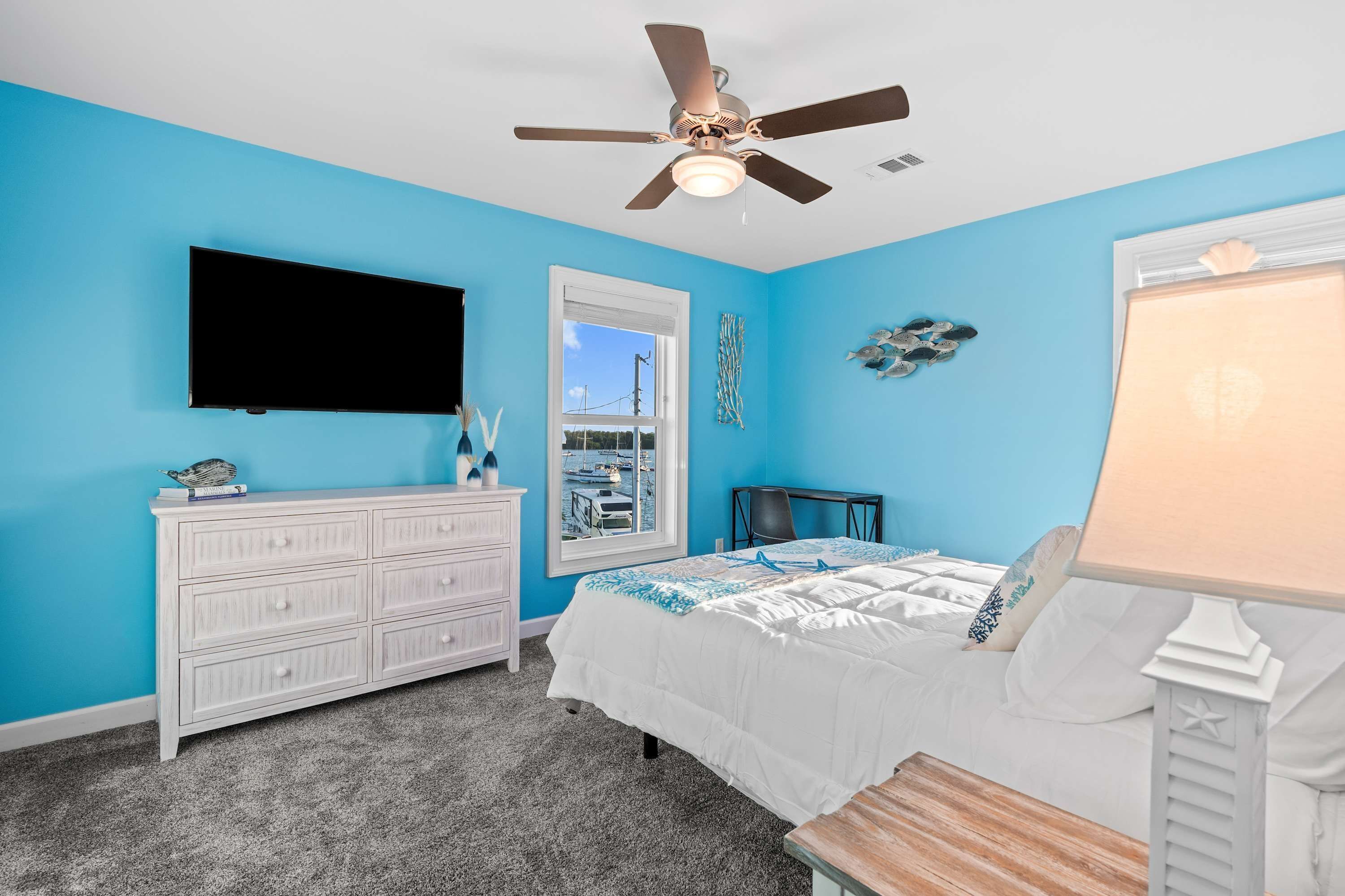 Een slaapkamer met een bed, een dressoir, een televisie, een plafondventilator, een raam en een lamp.