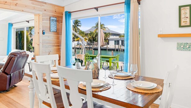 Un coin repas avec une table en bois et des chaises blanches, donnant sur une marina où l’on peut admirer des bateaux et des palmiers.