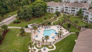 An aerial view of a resort with a central swimming pool, surrounded by palm trees and lounge chairs, with multi-story buildings in the background.