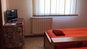 Una stanza piccola e luminosa con un letto, una televisione su un mobile colorato e un termosifone.
