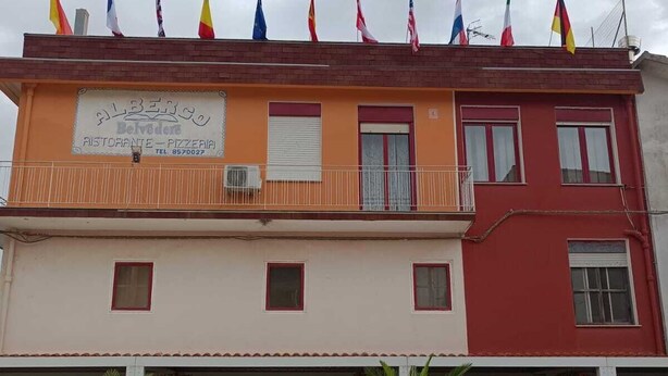 Un edificio con diverse bandiere, un cartello che indica 'Albergo Belvedere' come ristorante e pizzeria, e un ingresso chiuso da un cancello con vasi di piante.