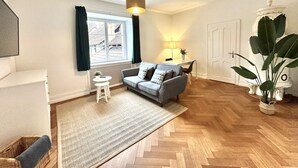 A modern living room with a grey sofa, a small white table, a large potted plant, and a window with curtains. - Business Apartment Solothurn (Solothurn)
