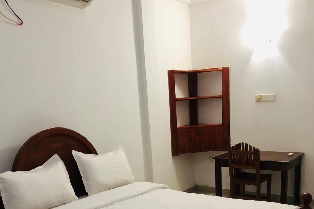 A neatly made bed with white bedding, a wooden headboard, a small wooden desk with a chair, and a wall-mounted shelf.