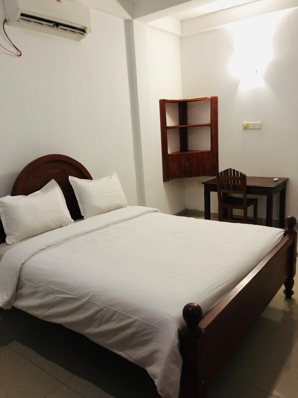 A neatly made bed with white bedding, a wooden headboard, a small wooden desk with a chair, and a wall-mounted shelf. - Viva la vida (Hikkaduwa)