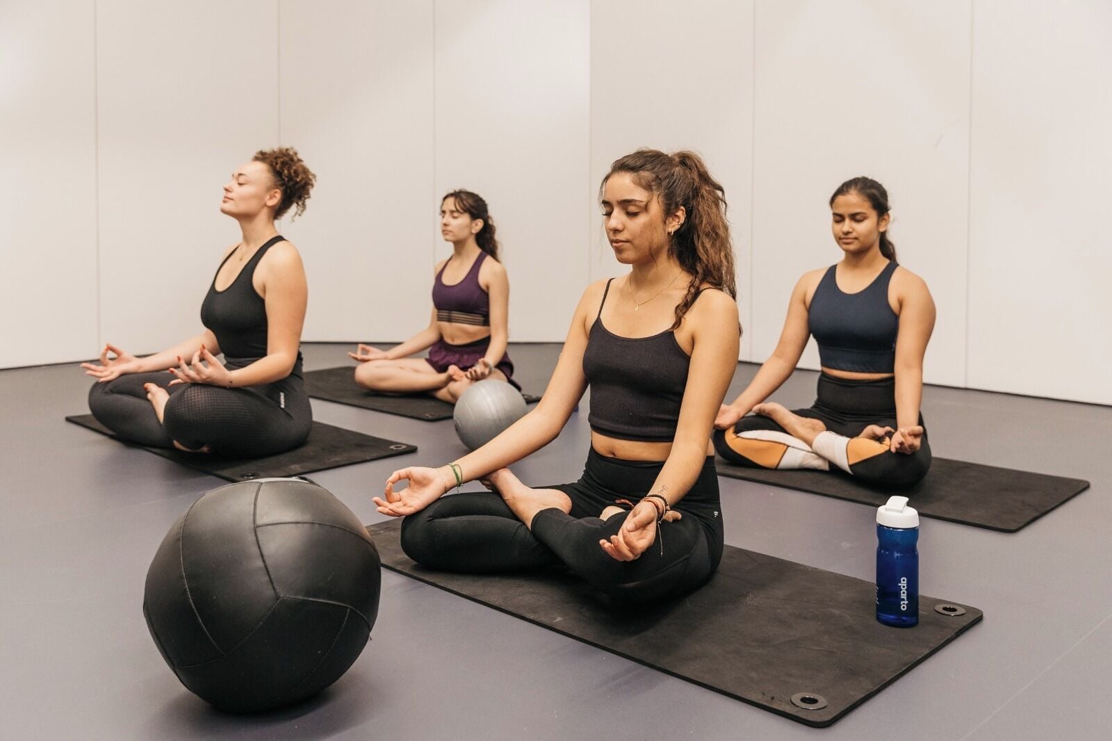 Four individuals seated on yoga mats in a room with a plain wall and a grey floor.