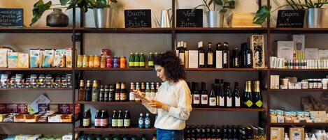 Una mujer con suéter blanco y jeans observa productos en estantes de una tienda, incluyendo botellas, cajas y frutas.