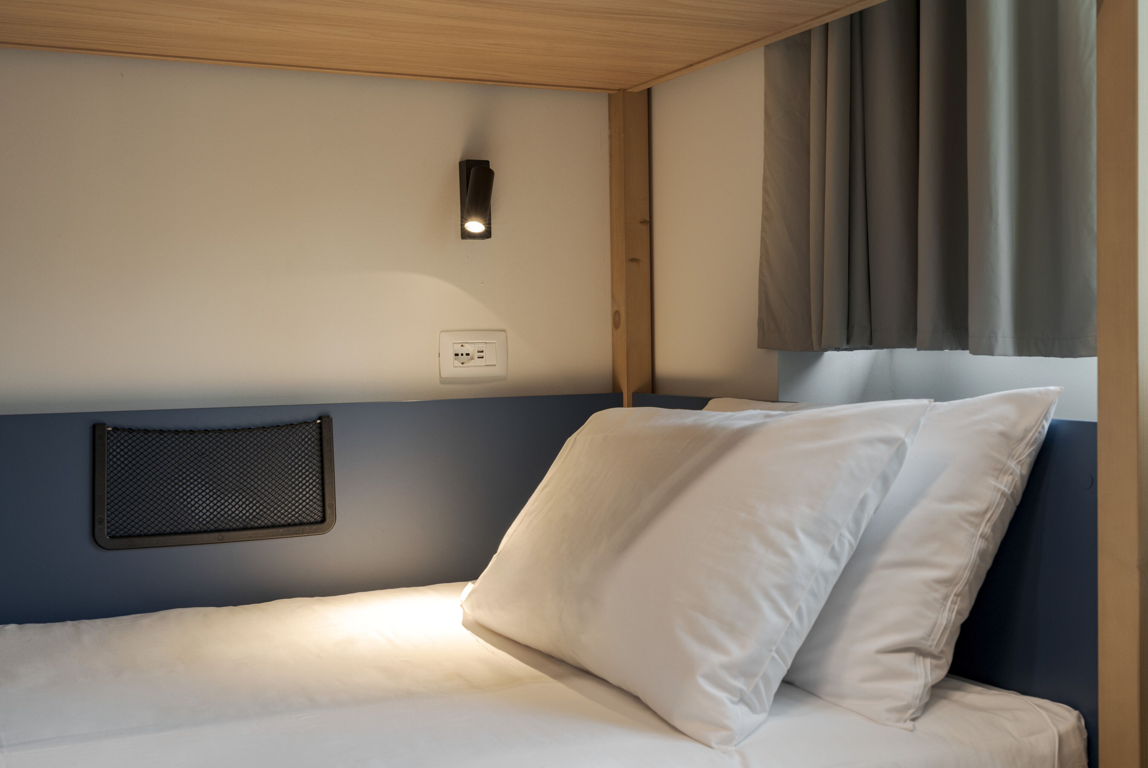 Una cama con ropa de cama blanca, una lámpara fijada a la pared y un enchufe.