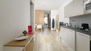 A modern kitchen with a microwave, coffee maker, and a sink. There is a wooden table with a white plate and a red box. A blue jacket hangs on a rack, and a wooden partition separates the kitchen from another room.