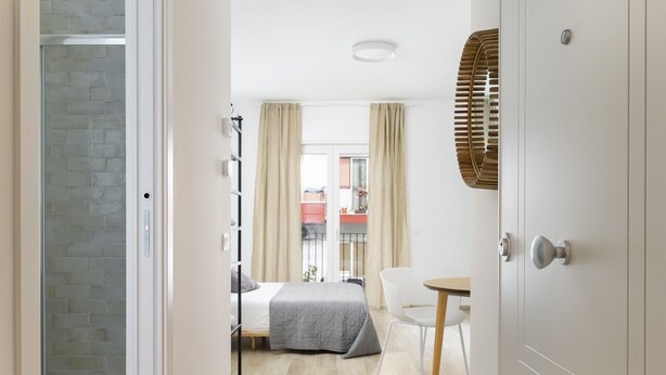 A hotel room with a bed, a chair, a small table, a window with curtains, and a door with a handle.