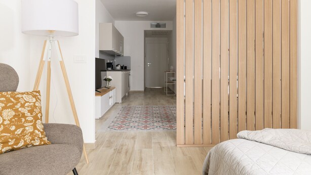 A modern, well-lit room with a sofa, a floor lamp, a bed, and a kitchen area in the background.
