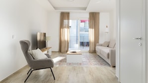 Ein modernes Wohnzimmer mit Sofa, Sesseln, Couchtisch, Fernseher und einem Balkon mit Vorhängen.