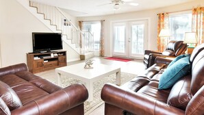 A living room with leather sofas, a white coffee table, a TV on a wooden stand, and a staircase. - Oceanfront 3 BR Condo with Shared Pool! Admiral's Quarters II 13 (Surfside Beach)