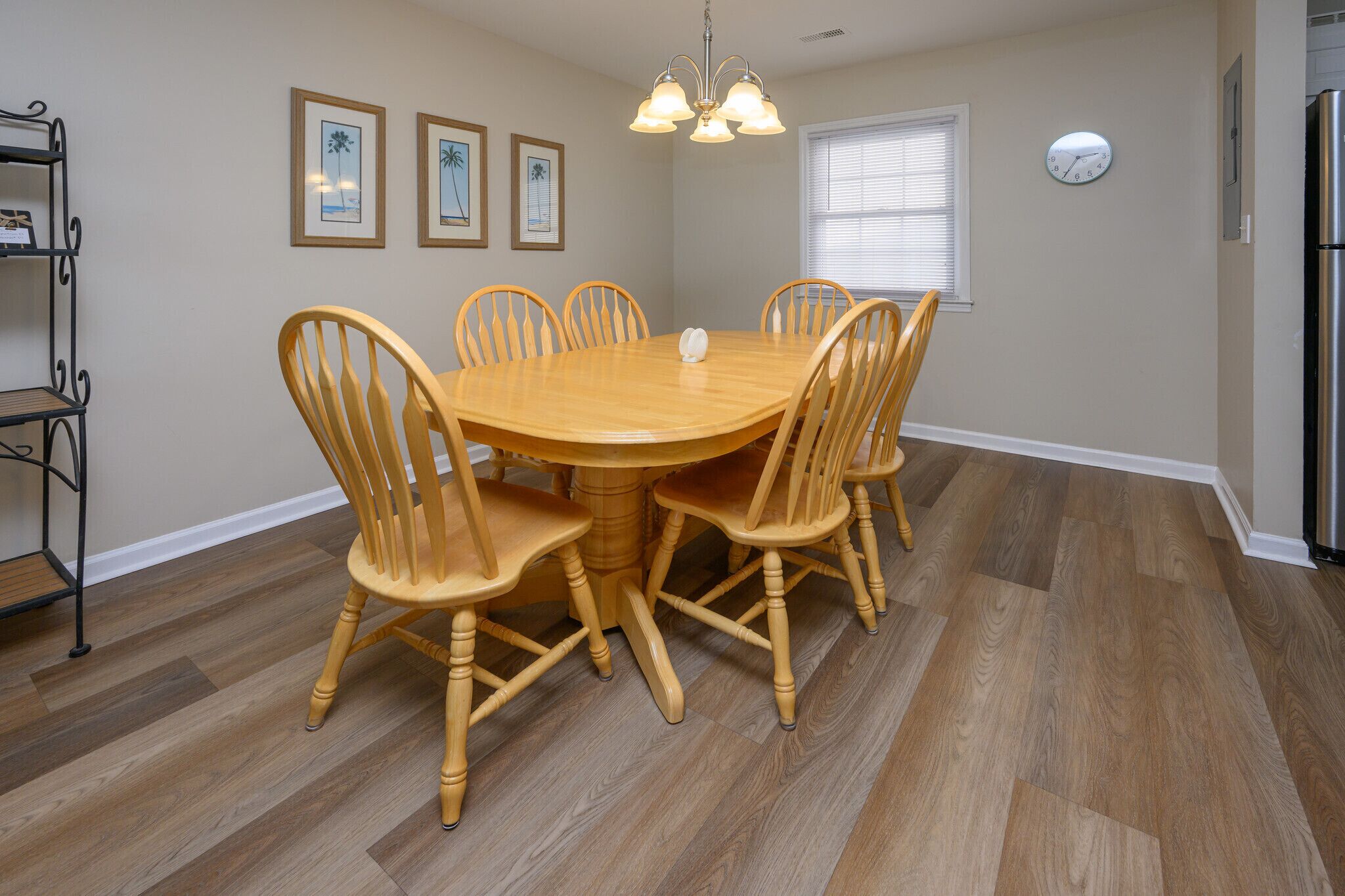 A dining area with a round wooden table surrounded by six chairs, a chandelier, framed pictures on the wall, and a clock.