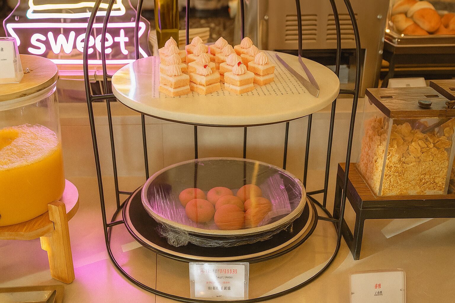 A buffet setup with various food items, including a neon sign reading "sweet," a glass display case with round pastries, and a container of orange juice.