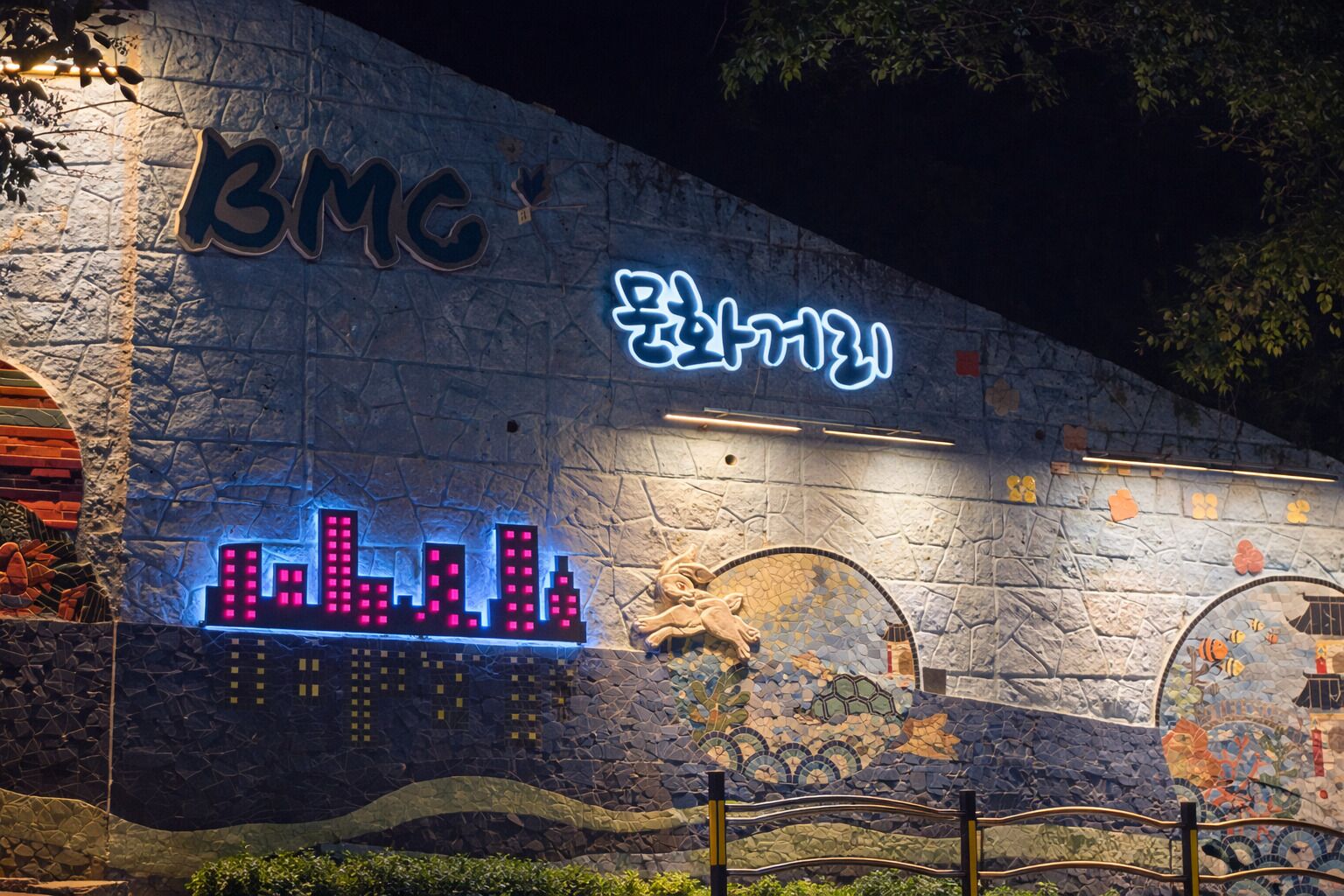 A building facade with a neon sign reading 'BMC' and a mosaic mural.
