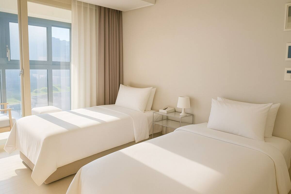 Una habitación de hotel con dos camas, una mesita de noche con lámpara y un ventanal con cortinas.