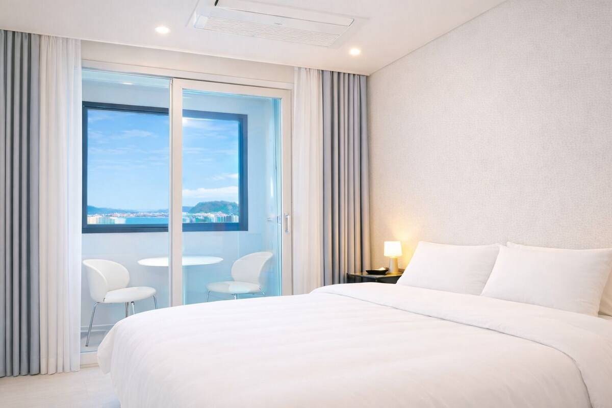 Una habitación de hotel con una cama grande, un balcón con dos sillas y vistas a una ciudad costera.