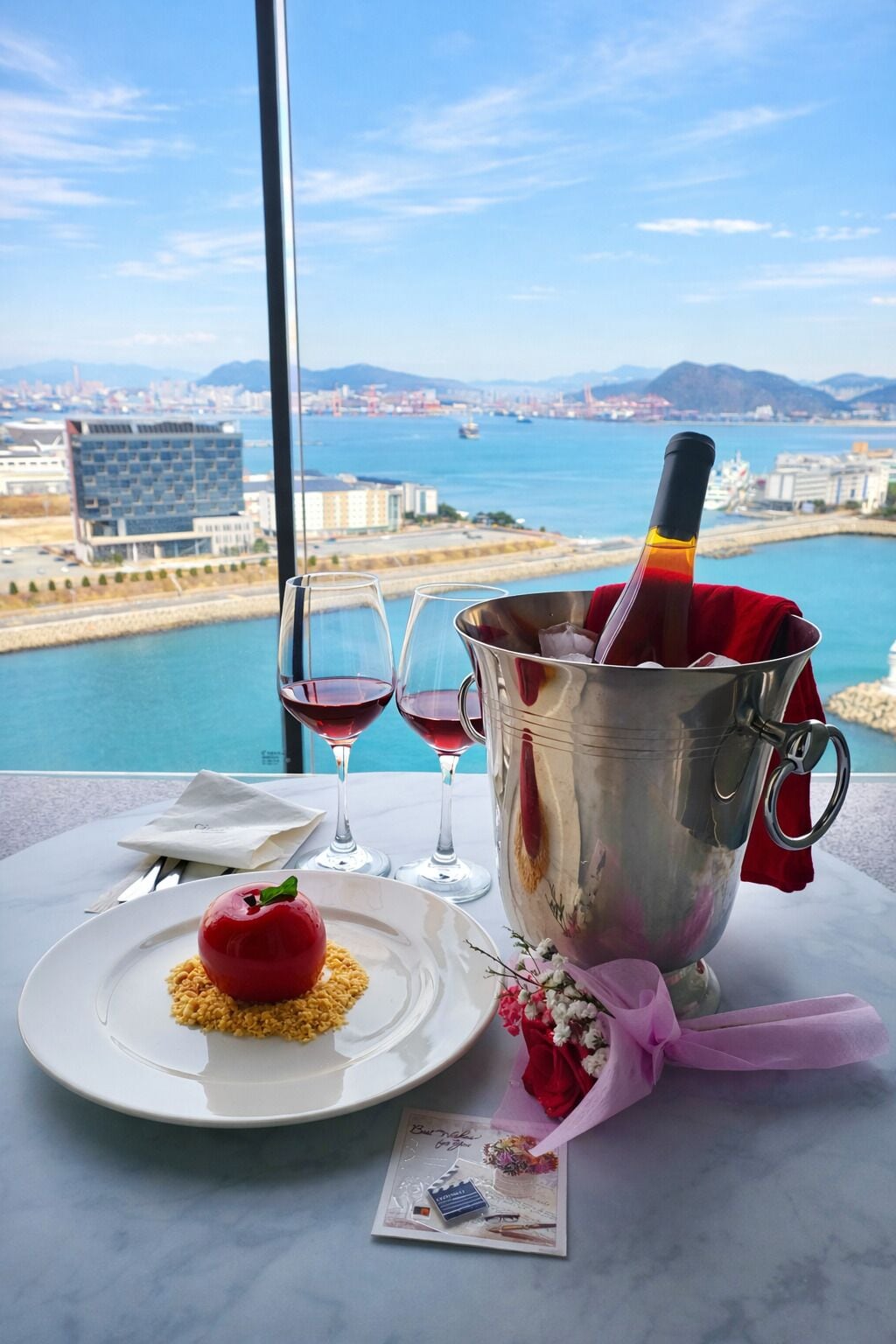 A table with a dessert, wine glasses, and a bottle of champagne in a bucket, overlooking a coastal cityscape.