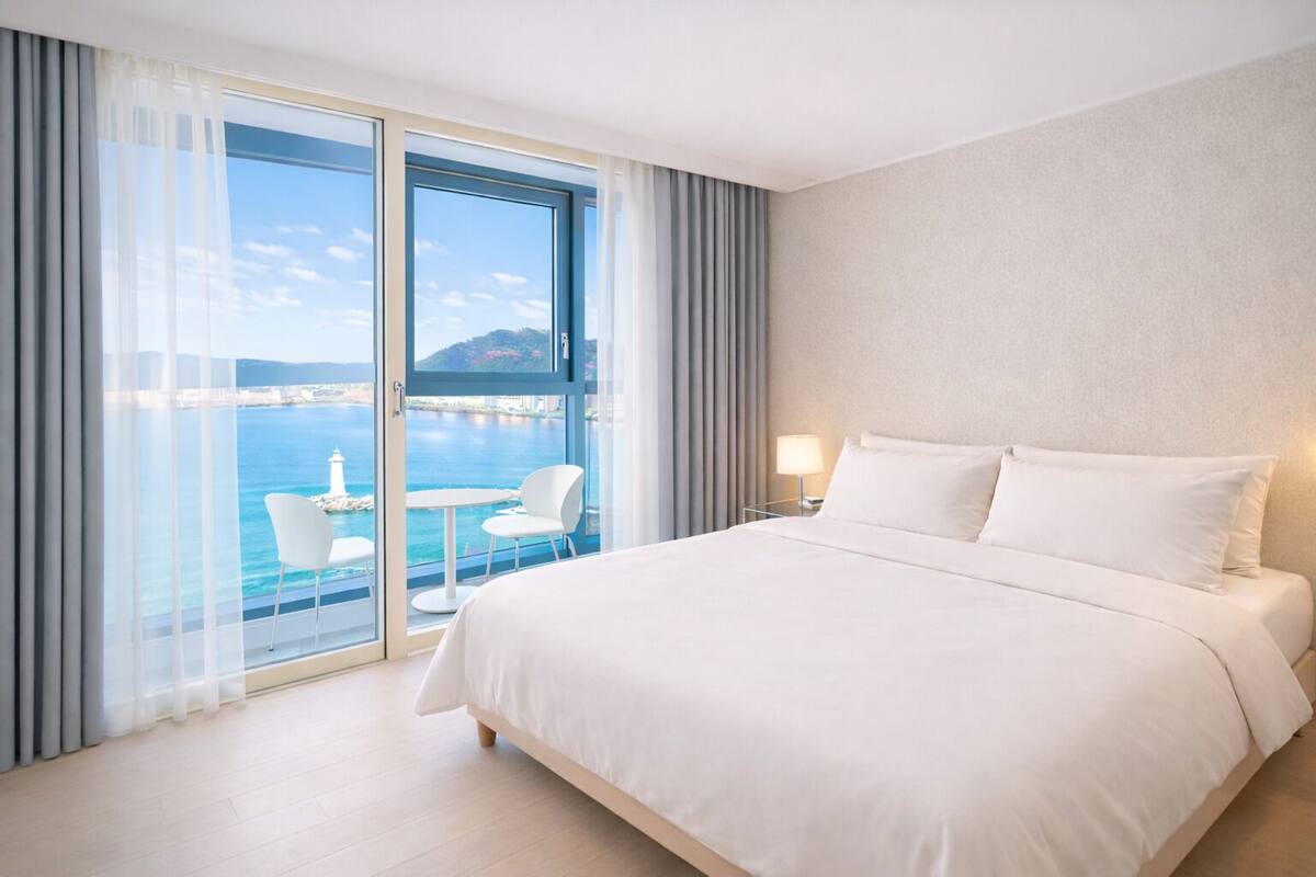 Una habitación de hotel con una cama grande, un balcón con vista al mar, y una pequeña mesa y silla.