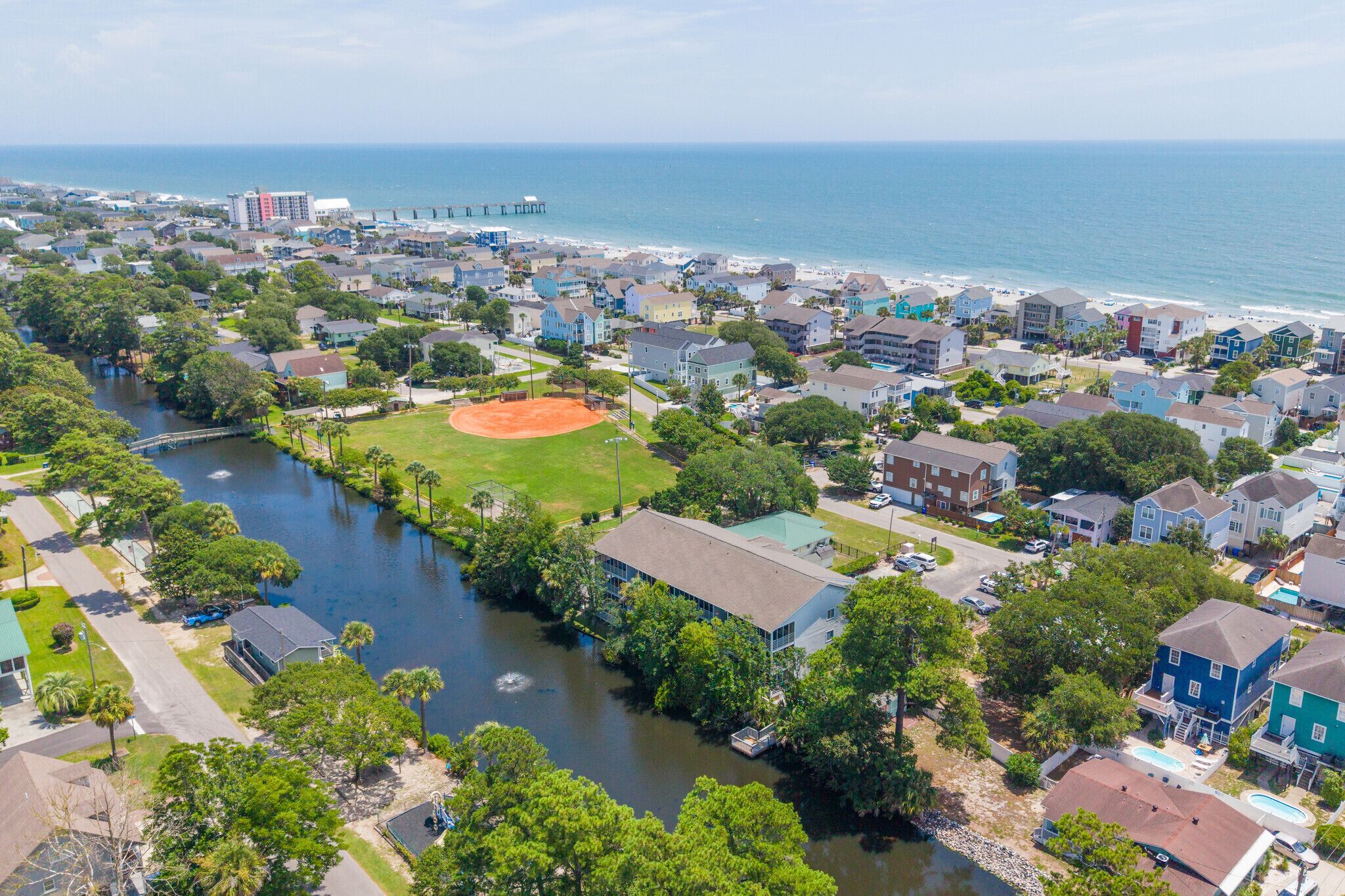 An aerial view of a coastal residential area with houses, a river, and a baseball field.