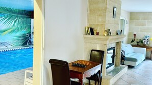 A living room with a dining table, chairs, a fireplace, and a view of a swimming pool. - VILLA, INDOOR POOL, NEAR SAINT-ÉMILION/ BORDEAUX, IDEAL FOR FAMILY. (Puynormand)