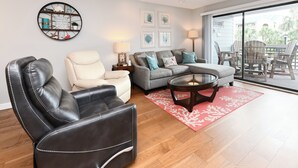 Un salon avec un fauteuil en cuir, un canapé et une table basse en verre. Une grande fenêtre offre une vue sur les palmiers.