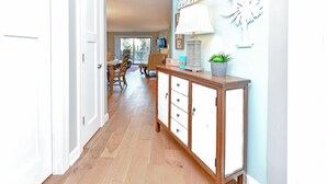 Un couloir avec un plancher en bois, une porte blanche, un meuble en bois avec des tiroirs, une table avec des chaises et un mur orné d’objets décoratifs.