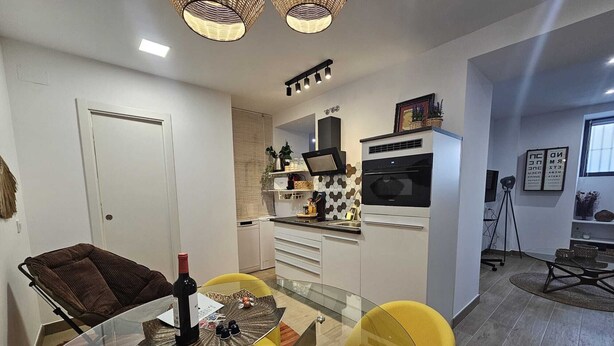 A modern kitchen with white cabinets, a black oven, and a microwave. There is a dining area with a glass table and yellow chairs. - The Gabriels Chamberi (Madrid)