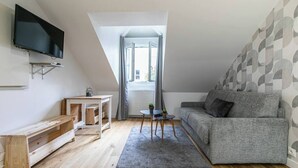 Ein modernes Loft-Apartment mit Schlafgalerie, einem grauen Sofa, einem kleinen Tisch, einem an der Wand montierten Fernseher und einem großen Fenster mit Vorhängen.
