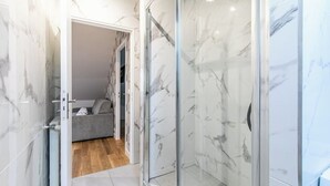 Ein modernes Badezimmer mit Marmorwänden, einer Duschkabine aus Glas und einem sichtbaren Schlafzimmer mit grauem Sofa.