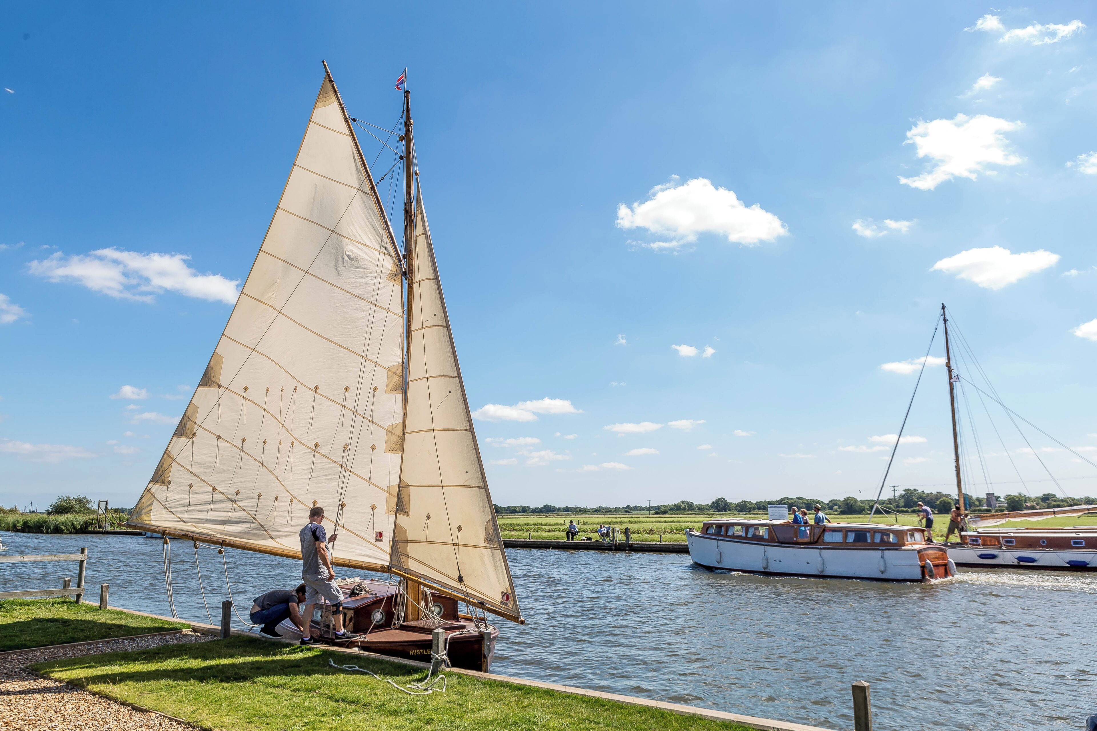A sailboat with a large sail on a calm river, docked near a grassy shore.