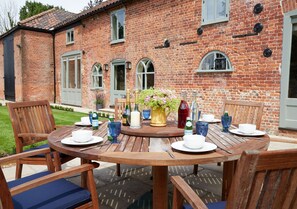 A wooden round table set for four with chairs, a brick building in the background, and a small garden area.