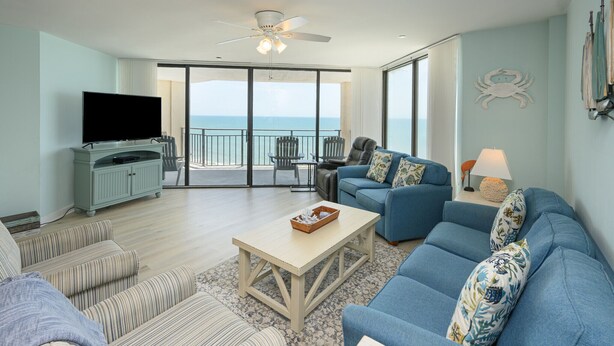 A spacious living room with a large TV, comfortable seating, a balcony with a view of the ocean, and a ceiling fan. - Oceanfront 3 BR Condo with a Shared Pool! Surf Master 807 (Garden City)