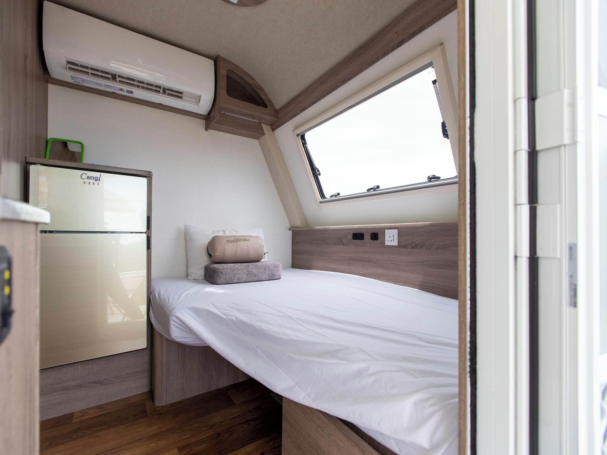 A compact RV interior with a kitchenette, a bed with a headboard, and a window providing natural light.